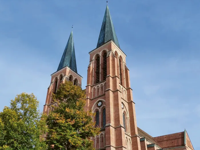© Elisabeth Metzler Zwei hohe, spitz zulaufende Kirchtürme aus rotem Backstein mit Bäumen im Vordergrund und blauem Himmel.