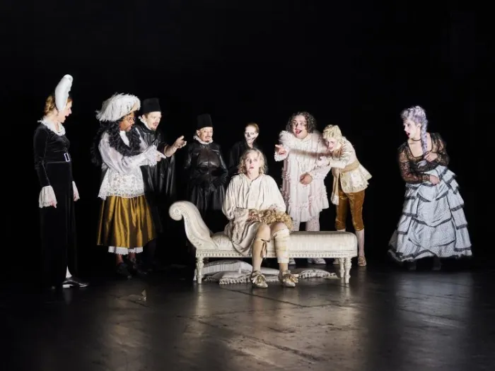 Szenenbild mit mehreren Personen in historischen Kostümen, eine Person sitzt auf einer Chaiselongue, andere stehen um sie herum auf einer dunklen Bühne.