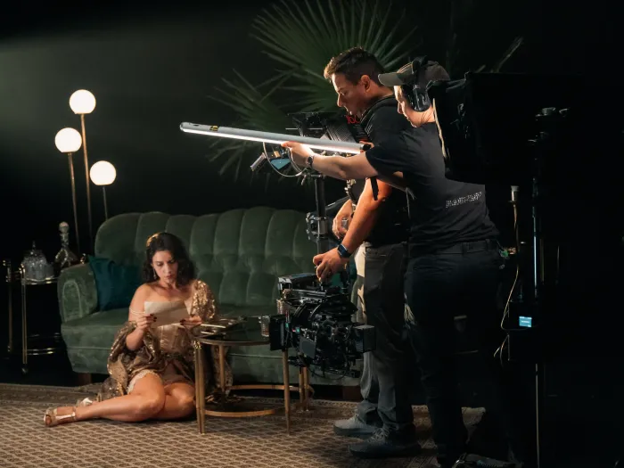 Frau in goldfarbenem Kleid sitzt auf Teppich vor grünem Sofa, zwei Männer bedienen Filmkamera und Lichttechnik.