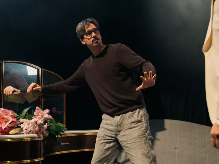 Mann in schwarzem Pullover und hellen Hosen steht mit ausgestreckter Hand vor einem Spiegel mit Blumen auf dem Tisch.