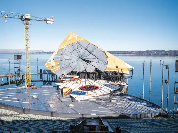 © Bregenzer Festspiele / Kristina Becker An open-air stage with rows of seats in front of a stage featuring a large, circular set piece and a crane by the lakeside under a clear sky.