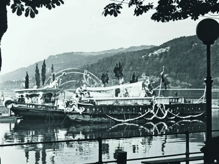 Black-and-white photo of a boat decorated with ornaments on a lake, with forested hills in the background and a tree in the foreground.
