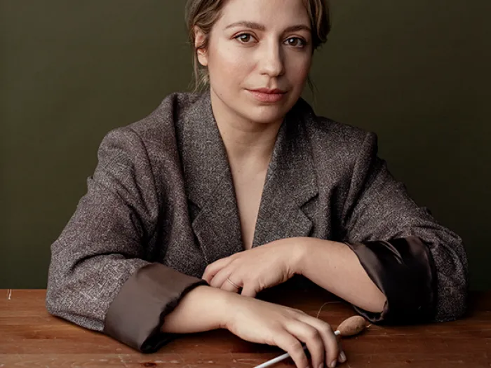 Dalia Stasevska © Sanna Lehto Person mit grauem Blazer sitzt an einem Holztisch und hält einen Dirigentenstab