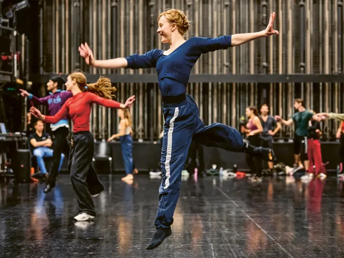 A dancer in blue training clothes is jumping with arms outstretched and one leg bent on a stage, with other dancers in the background.
