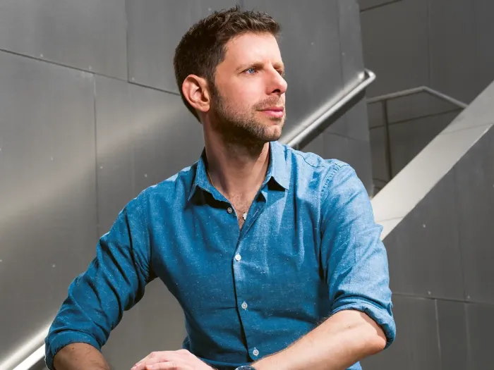 A man with a rolled-up shirt sleeve is sitting in front of a metallic wall with a staircase railing.