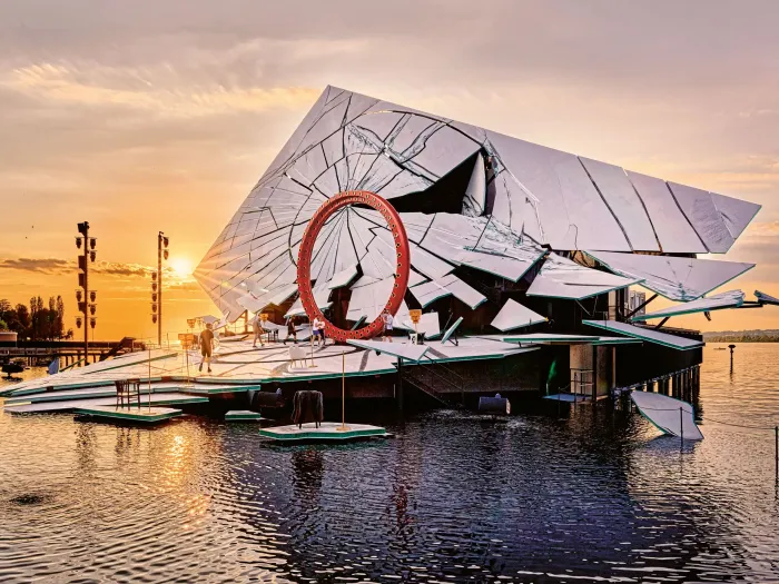 Seebühne mit großem roten Ring und spiegelnder, geometrisch gebrochener Fassade bei Sonnenuntergang auf einem See.