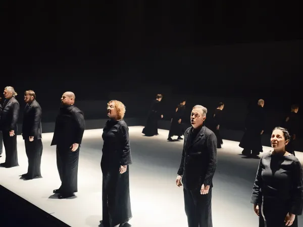 Mehrere Personen in schwarzer Kleidung stehen in einer Reihe auf einer Bühne mit dunklem Hintergrund und hellem Boden, sie blicken nach vorne und wirken konzentriert.