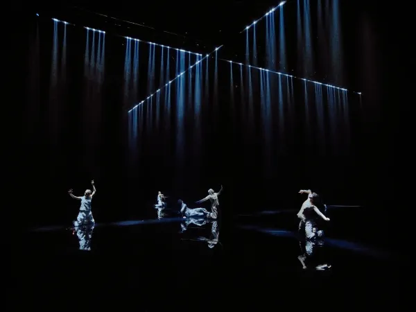 Vier Tänzer in fließenden Kostümen bewegen sich auf einer dunklen Bühne mit vertikalen und diagonalen blauen Lichtstrahlen, choreografiert von Tero Saarinen.
