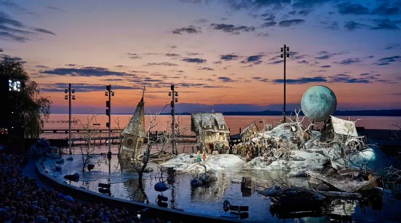 Seebühne 2024/25 "Der Freischütz: Ein winterliches Dorf, überflutet, es ragt ein Kirchturm aus dem Wasser, windschiefe Häuser, über dem Schneehügel schwebt ein Mond. Im Hintergrund geht die Sonne unter, der Himmel ist rot und blau. 