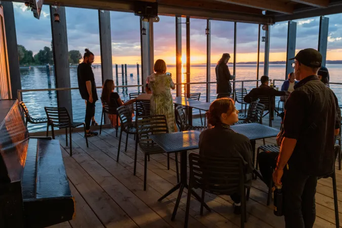 Menschen sitzen und stehen auf einer Terrasse mit Blick auf einen See bei Sonnenuntergang