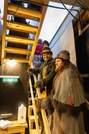 Drei Personen in historischen Kostümen mit Pelz und bunten Stoffen steigen eine Holztreppe in einem Theaterkeller hinauf