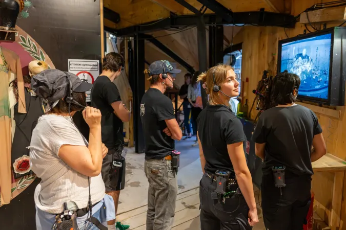 Mehrere Personen mit Headsets und Funkgeräten beobachten einen Monitor in einem Holzraum, vermutlich Backstage bei einer Theaterproduktion