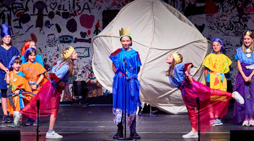 Szene aus "Meisterklasse B. Fassbaender", 2024 im Festspielhaus, Kinder in bunten Kostümen, mit Kronen oder Tüchern auf dem Kopf, im Hintergrund eine bemalte Wand und eine Konstruktion mit hellem Stoff überzogen 