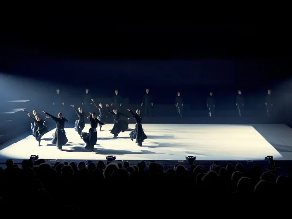 Tänzer in schwarzen Kostümen führen eine Choreografie auf einer hell beleuchteten Bühne vor dunklem Hintergrund auf