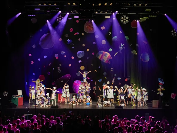 Kinder in bunten Kostümen führen eine Theateraufführung auf einer Bühne mit Weltraumdekoration und lila Bühnenlichtern auf