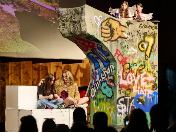 Zwei Mädchen sitzen im Schneidersitz auf einer Bühne neben einer bemalten Betonwand mit Graffiti und Schriftzügen wie "LOVE" und "HATE", auf der Betonwand sitzen zwei weitere Personen, im Vordergrund Publikum