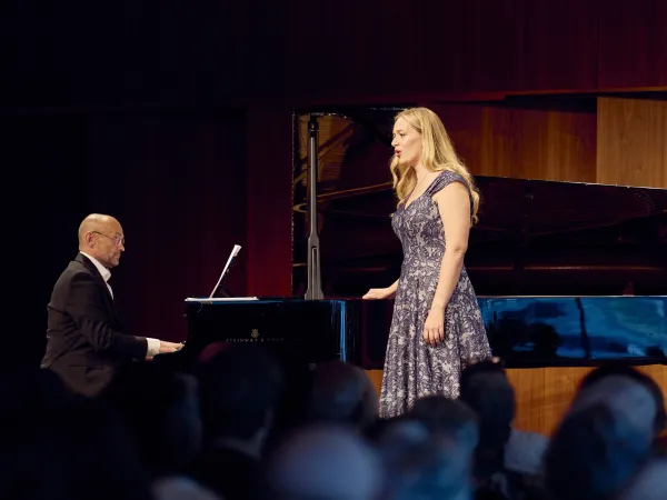 Frau in gemustertem Kleid singt neben einem Flügel, begleitet von einem Pianisten auf der Bühne