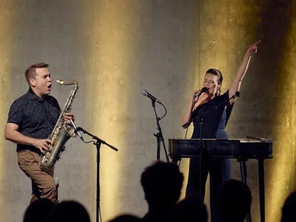 Musiker mit Saxophon und Sängerin mit Mikrofon auf Bühne vor Publikum, beleuchtet von warmem Licht