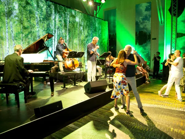 Live-Band mit Klavier, Kontrabass und Sänger spielt auf Bühne vor tanzenden Paaren in einem Saal mit grünem Licht und Waldkulisse