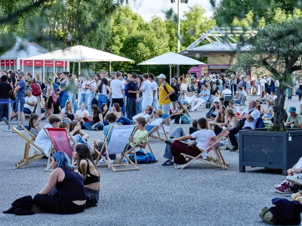 Menschen sitzen und stehen beim Festspielplatz mit Liegestühlen und Sonnenschirmen, umgeben von Bäumen