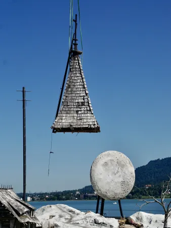 Schindelgedecktes, spitz zulaufendes Dachteil wird an einem Kranhaken über einem See gehoben, daneben eine große runde Skulptur mit Mondoberfläche.
