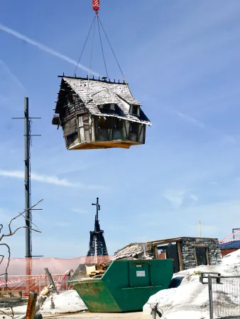 Altes, kleines Holzhaus mit schneebedecktem Dach wird von Kranhaken in der Luft über einer Baustelle mit Containern und Bauzäunen gehoben.