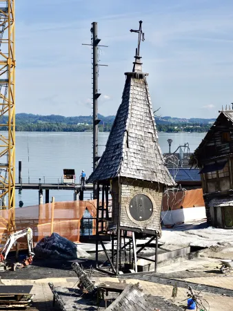 Alter Uhrenturm mit schiefergedecktem Dach und großem Zifferblatt, umgeben von Baustelle am Seeufer mit Kran und Bauzaun