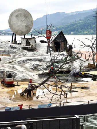 Zwei Personen mit roten Helmen bewegen einen großen kahlen Baumstamm auf einer Bühne mit Kulissen von Hütten und einem großen Mond im Hintergrund.