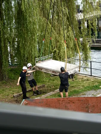 Drei Männer mit Helmen heben eine große Betonplatte mit einem Kran an, im Hintergrund ein Fluss und Weidenzweige.