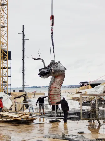 Große Skulptur eines Schlangenkopfs mit rotem Hals wird an einer Baustelle von einem Kran gehoben, mehrere Personen beobachten.