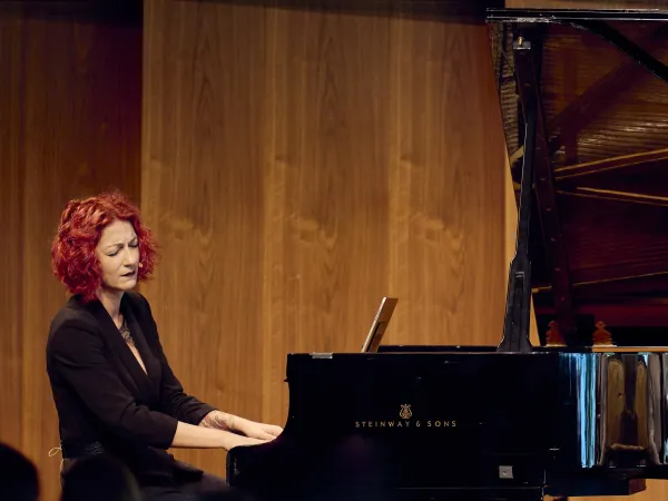 Frau mit rotem, lockigem Haar sitzt an einem schwarzen Steinway & Sons Klavier. Sie trägt ein schwarzes Outfit und spielt. Im Hintergrund ist ein offenes Klavier sichtbar.