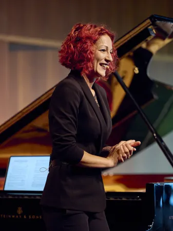 Frau mit rotem, lockigem Haar in schwarzem Blazer, steht neben einem Klavier. Im Hintergrund ist ein offenes Klavier und Notenblätter sichtbar.