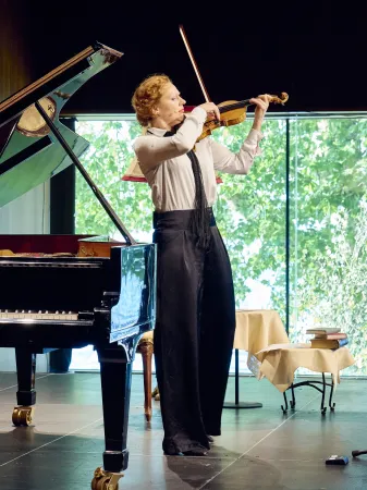 Eine Person spielt Violine, gekleidet in ein weißes Hemd und schwarze Hose. Im Hintergrund steht ein Klavier und ein Tisch mit Büchern.