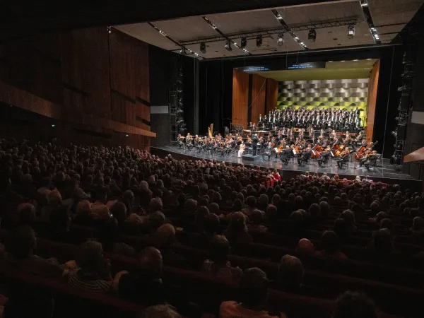 Ein Orchesterkonzert mit einem großen Publikum. Im Vordergrund steht ein Dirigent vor einem Orchester mit Streichern und Bläsern. Im Hintergrund ist ein Chor zu sehen.