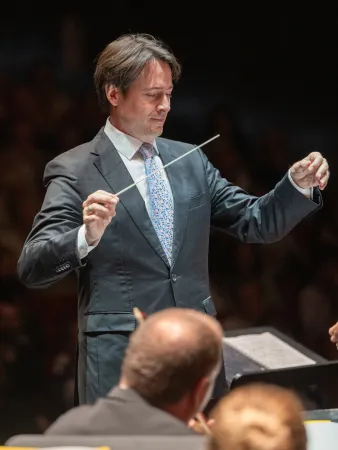 Dirigent Leo McFall dirigiert mit Taktstock vor schwarzem Hintergrund und trägt einen Grauen Anzug und eine hellblaue Krawatte