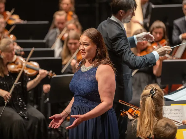 Sängerin in dunkelblauem Kleid mit braunen Haaren. Im Hintergrund das Orchester und der Dirigent mit schwarzem Anzug