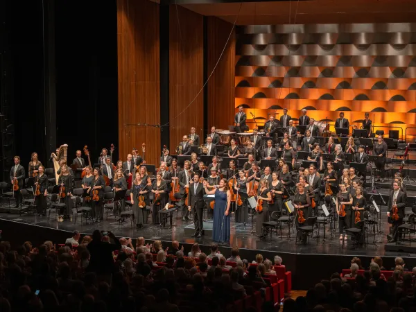 Gesamtes Orchester auf der Bühne mit orange beleuchtetem Hintergrund. Die beiden Solisten verbeugen sich und das Orchester steht.