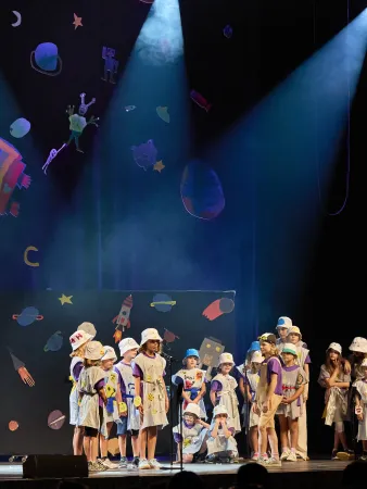 Eine Gruppe von Kindern in bunten Kostümen steht auf einer Bühne, umgeben von einem farbenfrohen, bemalten Hintergrund mit Weltraummotiven. Zwei Scheinwerfer strahlen auf die Gruppe.
