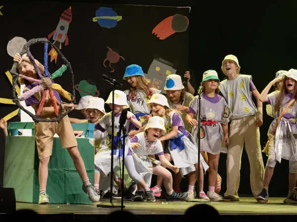Eine Gruppe von Kindern auf einer Bühne in Kostümen, die verschiedene Farben und Muster aufweisen. Ein Kind hält ein großes, buntes Rad. Im Hintergrund sind Zeichnungen von Raketen und anderen Objekten.