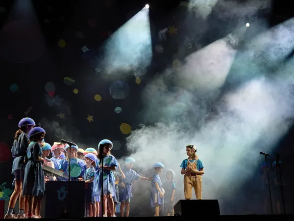 Eine Gruppe von Kindern in blauen Kostümen steht auf einer Bühne. Ein Kind in einem beigen Outfit steht vorne und schaut zur Gruppe. Nebel und Scheinwerferlicht sind sichtbar.