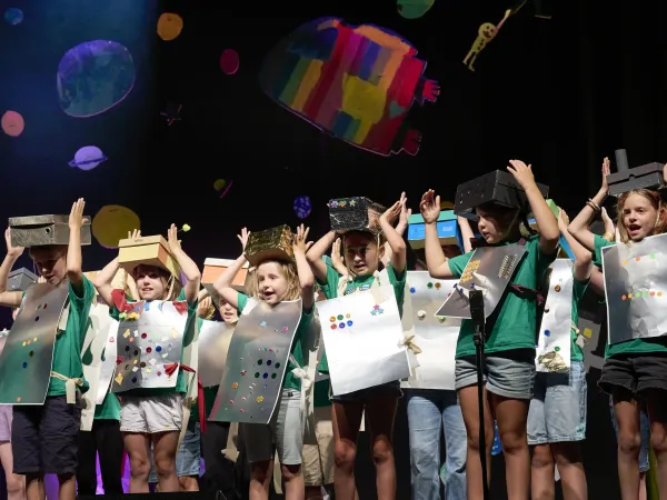 Gruppe von Jugendlichen in grünen T-Shirts, die bunte, selbstgemachte Kostüme aus Karton tragen. Sie halten ihre Arme hoch und stehen auf einer Bühne.