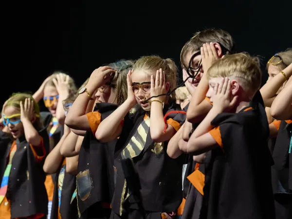 Gruppe von Kindern auf einer Bühne, die schwarze Umhänge mit orangefarbenen Akzenten tragen. Einige Kinder halten sich die Hände an den Kopf.