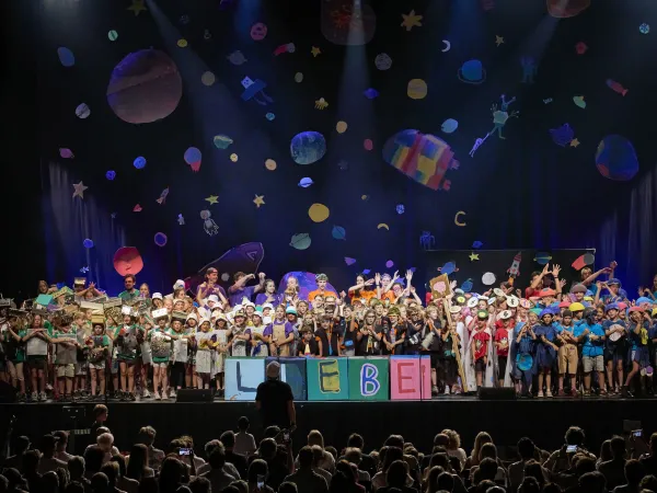 Eine große Gruppe von Kindern steht auf einer Bühne, trägt bunte Kostüme und hält ein Schild mit dem Wort 'LIEBE'. Der Hintergrund zeigt farbenfrohe, gemalte Planeten und Sterne.