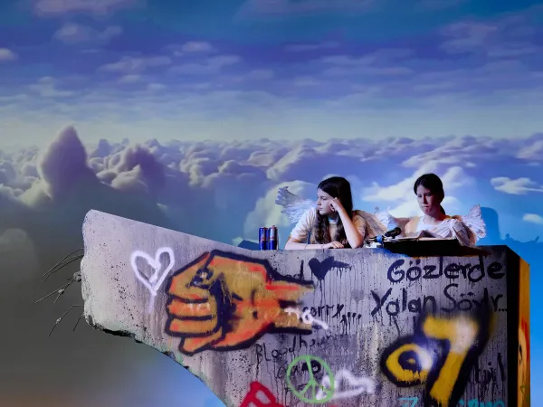 Zwei Personen mit Flügeln sitzen auf einer grauen Wand mit Graffiti. Die Wand zeigt eine große, bunte Hand und verschiedene Symbole wie Herzen und Frieden. Im Hintergrund sind Wolken zu sehen.