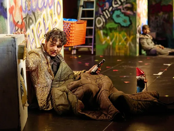 Eine Person sitzt auf dem Boden, eingehüllt in eine dicke, braune Decke. Sie hält eine Flasche in der Hand. Im Hintergrund sind bunte Graffiti und eine Leiter sichtbar. Ein kleiner Gnom aus Holz steht neben der Person.