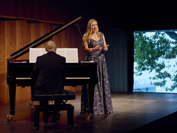 Eine Sängerin in einem langen, blauen Abendkleid steht auf einer Bühne und singt. Ein Pianist sitzt mit dem Rücken zur Kamera an einem schwarzen Klavier.