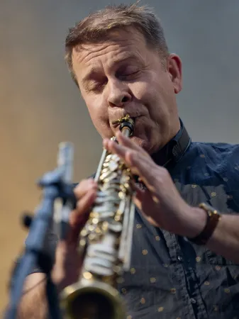 Ein Musiker spielt eine Flöte. Er trägt ein schwarzes Hemd mit goldenen Punkten und hat eine Armbanduhr am Handgelenk.
