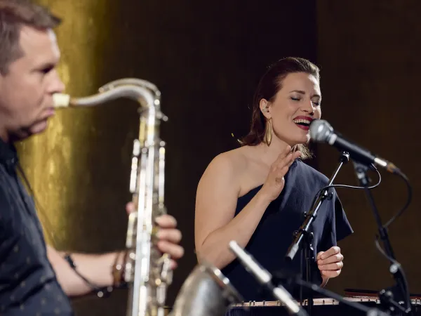 Eine Frau mit schulterfreier, schwarzer Bluse steht am Mikrofon und gestikuliert. Im Vordergrund ist ein Saxophonspieler zu sehen.