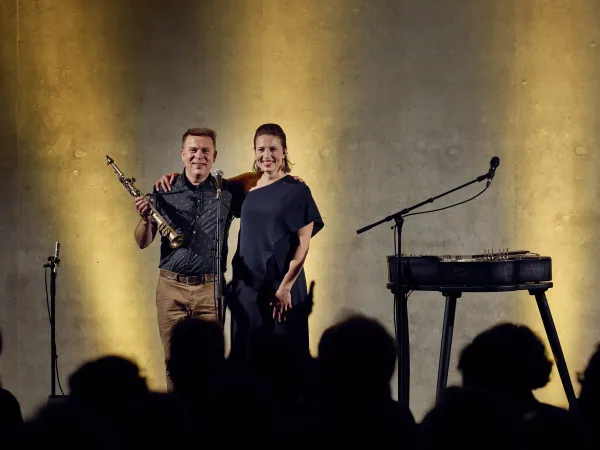Ein Mann hält ein Saxophon in der linken Hand, steht neben einer Frau in einem schwarzen Kleid. Im Hintergrund sind Mikrofone und ein Tisch mit Musikinstrumenten sichtbar.