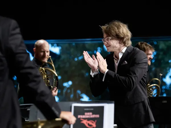 © Bregenzer Festspiele / Anja Koehler Ein Mann in einem schwarzen Anzug mit einem weißen Hemd applaudiert. Im Hintergrund sind Musiker mit Hörnern zu sehen. Auf einem Notenständer steht 'DAS PHILHARMONIKER'.